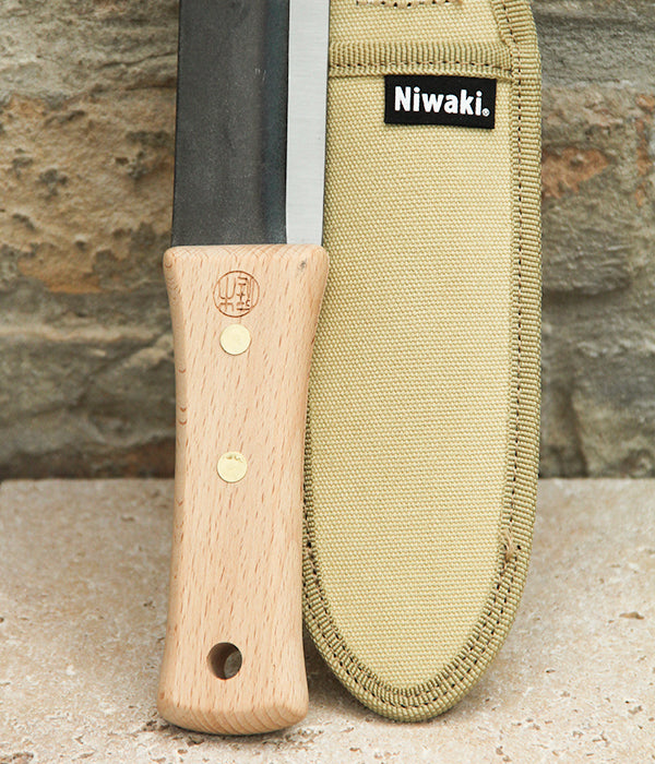 Das Niwaki | „Hori Hori“ – ein multifunktionales Pflanzmesser mit Schutzhülle und robustem Holzgriff – liegt neben seinem beigen Etui mit Niwaki-Logo – elegant auf einem Stein vor einer Backsteinmauer präsentiert und perfekt geeignet für all Ihre Gartenarbeiten.