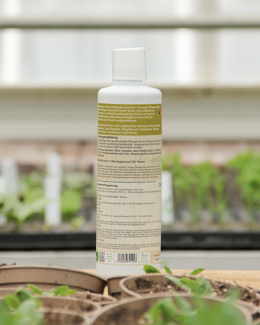 Eine weiße Kunststoffflasche von AMN | 'Optiferrum' Organischer Spurennährstoffdünger und Bodenvitalstoff 0,5 l steht auf einer Holzfläche zwischen Sämlingstöpfen in einem Gewächshaus. Das Etikett in deutscher Sprache und die Rückseite der Flasche sind sichtbar.