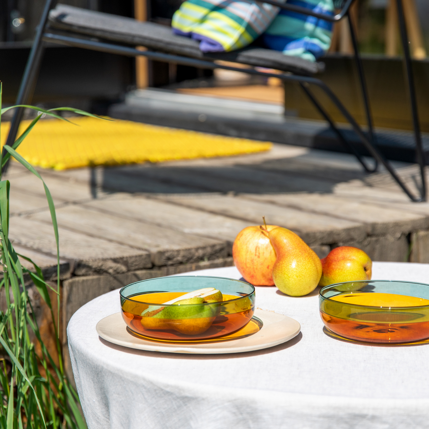 Zwei 'Amber' Glasschälchen aus Borosilikatglas aus dem Remember 2er Set der Königlichen Gartenakademie halten Obst auf einem weiß gedeckten Tisch im Freien; daneben ruhen Birnen auf der Terrasse mit Blick auf einen Liegestuhl und Handtücher.
