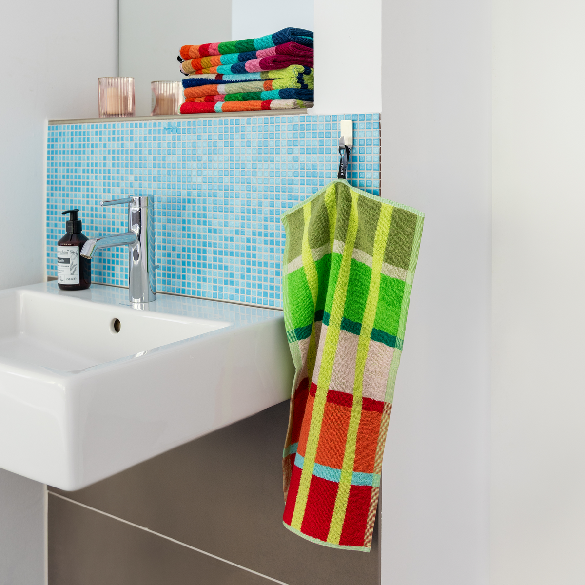 Ein Waschbecken mit einer verchromten Armatur, einer blauen Mosaikfliesen-Rückwand, einem Stapel bunter Badetücher aus hochwertiger Baumwolle, einem Seifenspender und dem „Kaido“-2er-Set Gästehandtücher von Remember in grün-orangefarbenem Karomuster, das an einem Haken hängt.