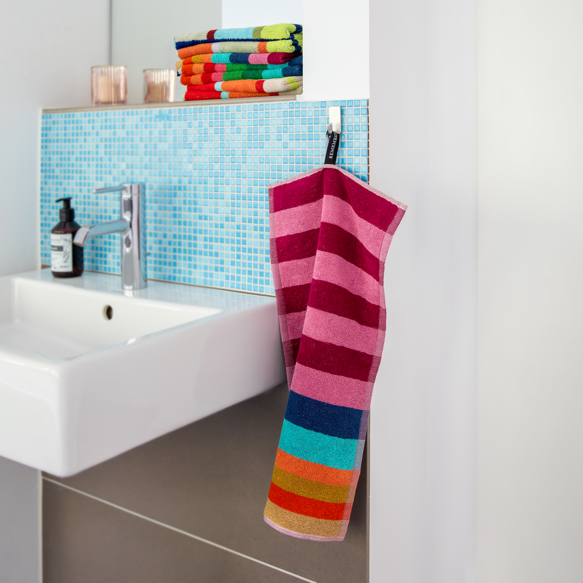 Ein „Remember“-Gästehandtuch „Alva“ aus dem 2-teiligen Set hängt an einem Haken neben einem Waschbecken mit einer blauen Mosaikfliesen-Rückwand. Auf der Waschbeckenkante liegen passend gefaltete Handtücher aus weicher 500 g/m²-Baumwolle von Remember sowie eine Seifenablage.