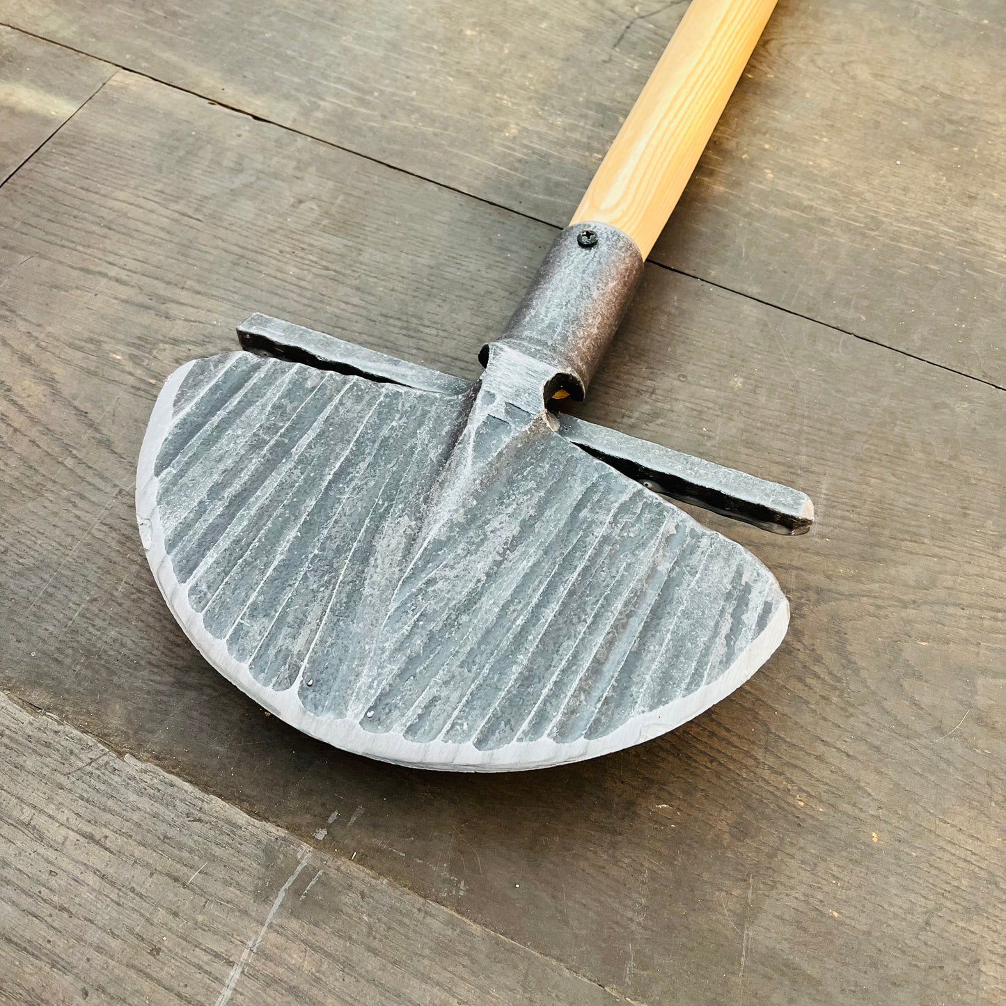 Eine Nahaufnahme des Krumpholz Rasenkantenstechers 85cm von Krumpholz mit einem Holzgriff und einer breiten, geriffelten, halbrunden Metallklinge, die auf einer Holzunterlage liegt.