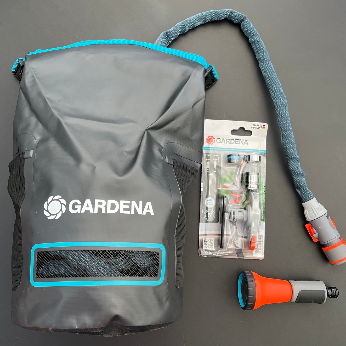 Ein Gardena Textilschlauch Liano™ Xtreme 1/2" Balkon und Terrasse, ein Gardena Zubehörset in der Verpackung und eine orange-graue Sprühdüse, alles auf einer dunklen Fläche ausgestellt.