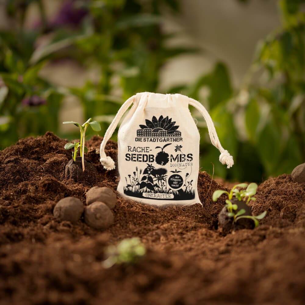 Ein kleiner Stadtgärtner Jutebeutel mit der Aufschrift "Samenbomben 'Rachebombs Beutel' Unkraut" steht in der Erde, umgeben von Samenkugeln und Sämlingen, mit verschwommenen grünen Pflanzen im Hintergrund.