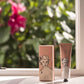 Eine Tube und eine Schachtel der "Kirschblüte"-Handcreme von Archivist Gallery mit einem illustrierten Frauengesicht auf der Verpackung liegen auf einer Fensterbank, vor der verwischte rosa Blumen und Grünpflanzen stehen.