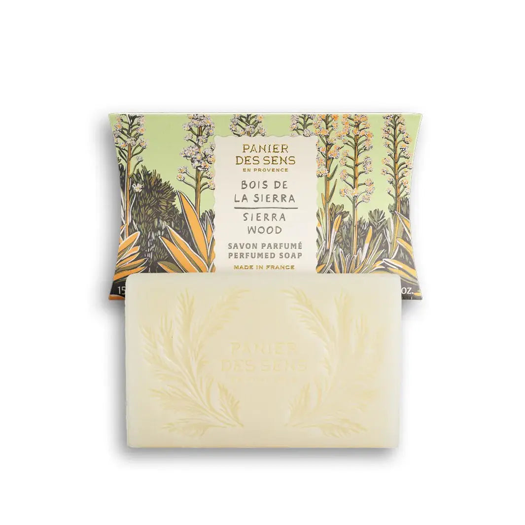 Ein Barren Panier Des Sens | Duftseife Sierraholz 150 g, geprägt mit einem botanischen Design, steht vor seiner mit Blumen illustrierten Verpackung und verströmt einen holzig-würzigen Duft.
