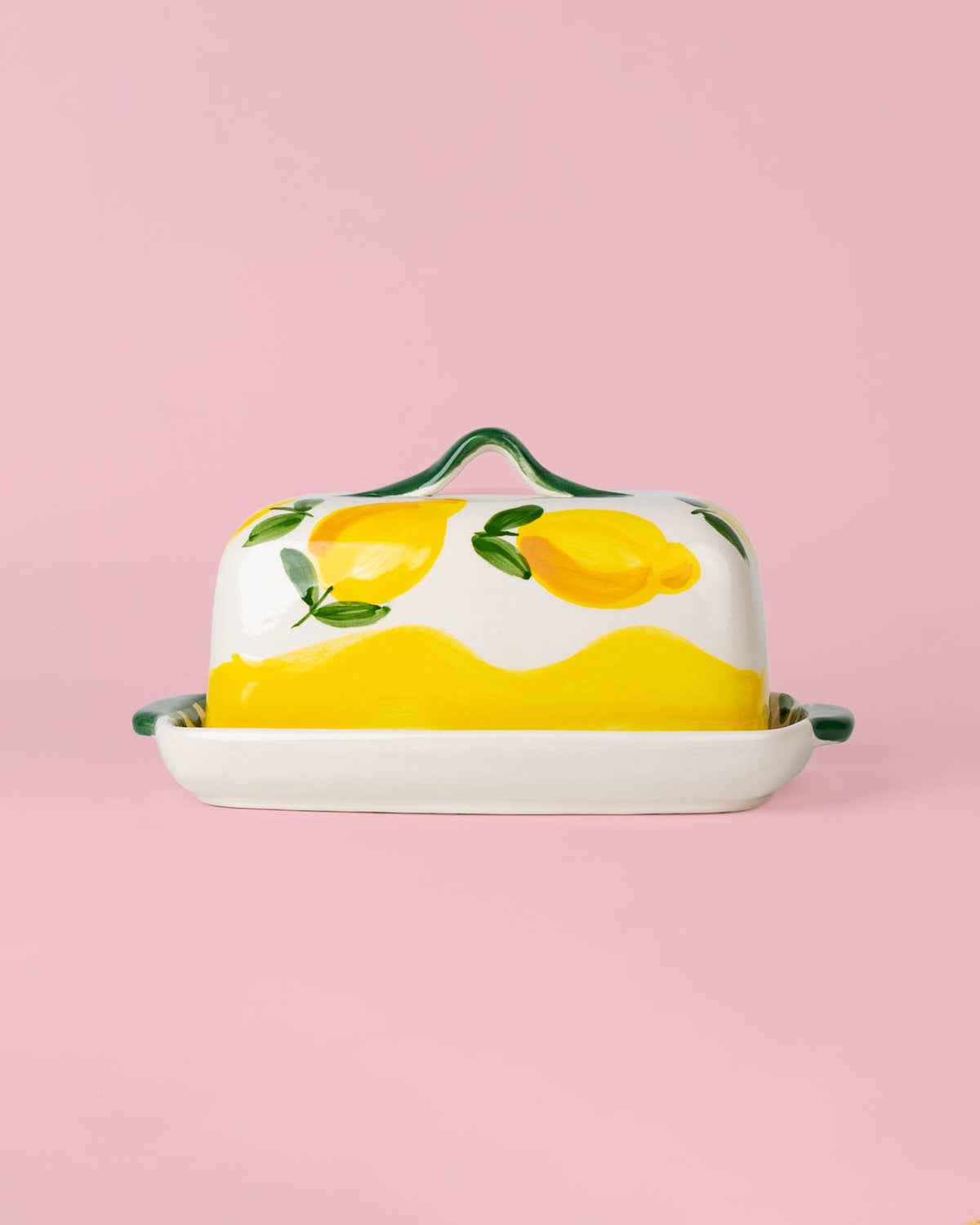 Die handgefertigte Keramik-Butterdose „Limone“ in Gelb und Weiß von Alfar Tierra Cocida, kunstvoll verziert mit gelben Zitronen und grünen Blättern, präsentiert sich vor einem hellrosa Hintergrund – ein stilvoller Akzent für mediterrane Tischkultur.