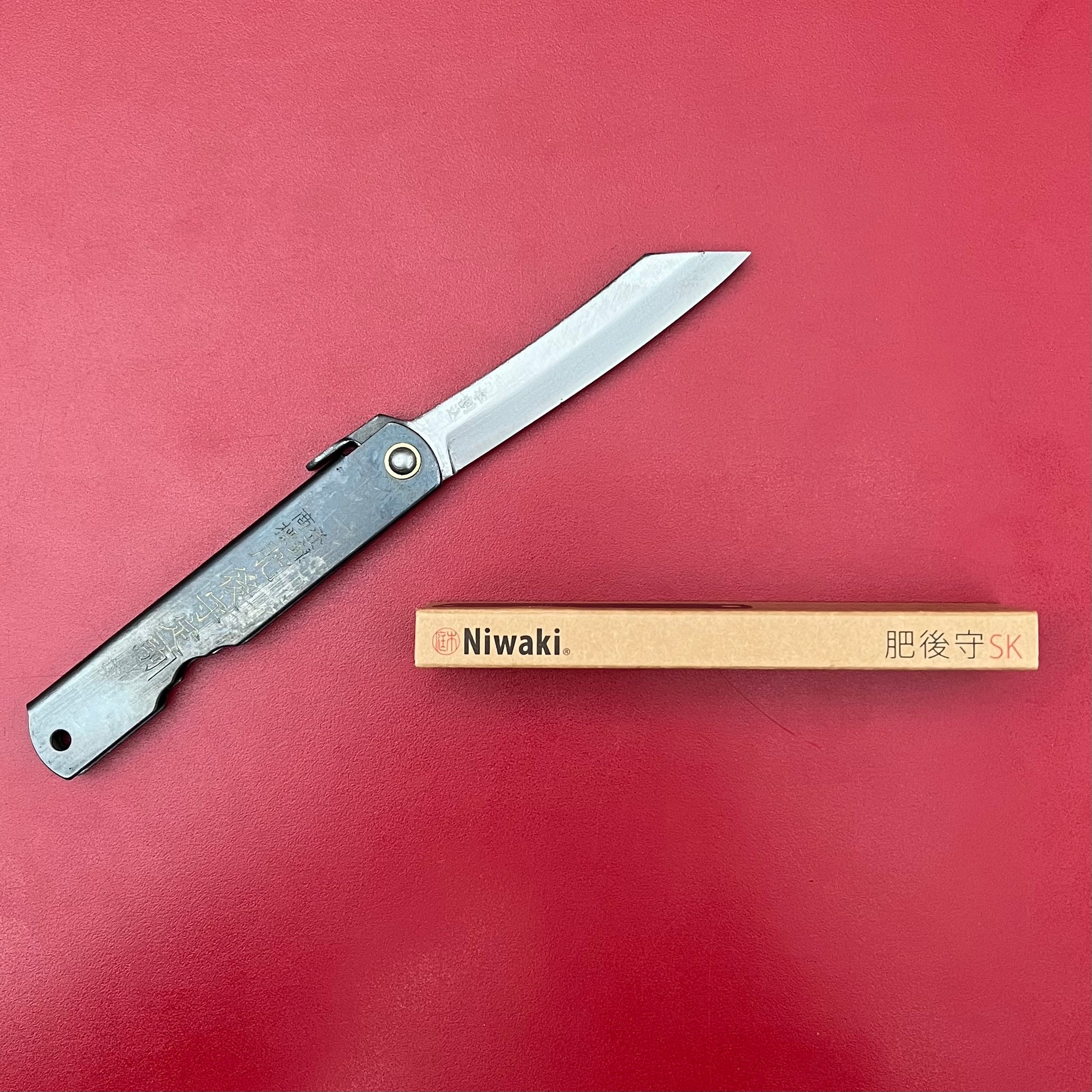 Das Niwaki | 'SK Higonokami Folding Knife' Klappmesser von Niwaki mit Metallgriff und gravierter Klinge liegt neben der rechteckigen Verpackungsbox auf rotem Untergrund.