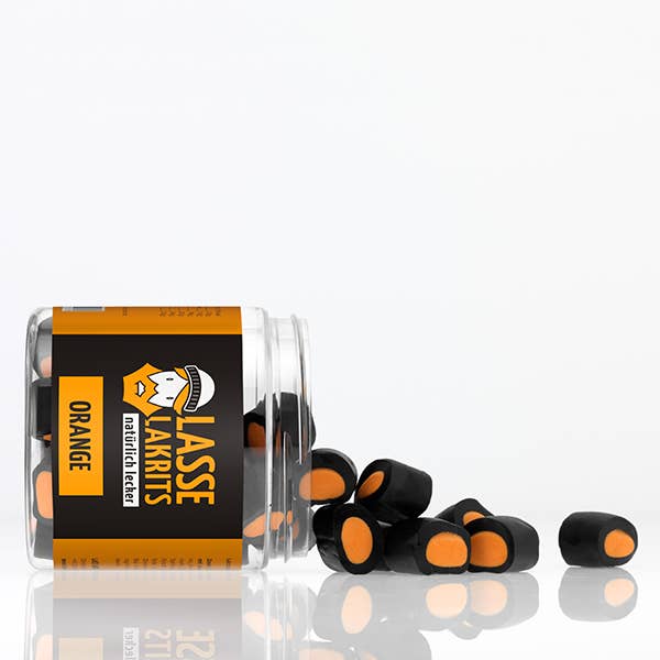Ein LASSE LAKRITS | Dose Lakritz Orange Glas von LASSE LAKRITS liegt auf der Seite und verschüttet fruchtige Lakritze und orangefarbene Bonbonstücke ohne Palmöl auf eine weiße Fläche.
