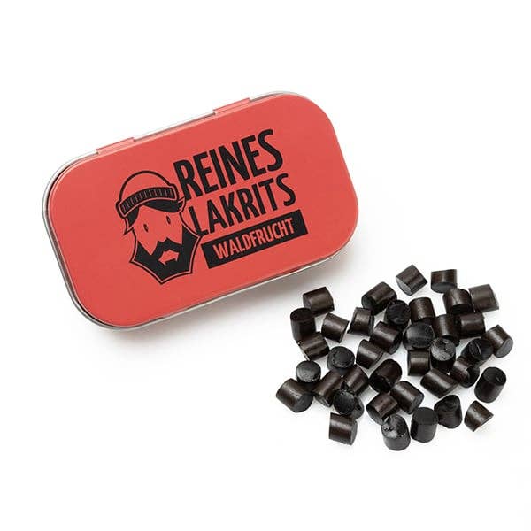 Eine rote LASSE LAKRITS-Dose mit der Aufschrift "Dose Reines Lakritz Waldfrucht", auf der ein bärtiges Gesicht abgebildet ist, steht neben kleinen schwarzen zuckerfreien Lakritzbonbons, die auf einer weißen Fläche verstreut sind.