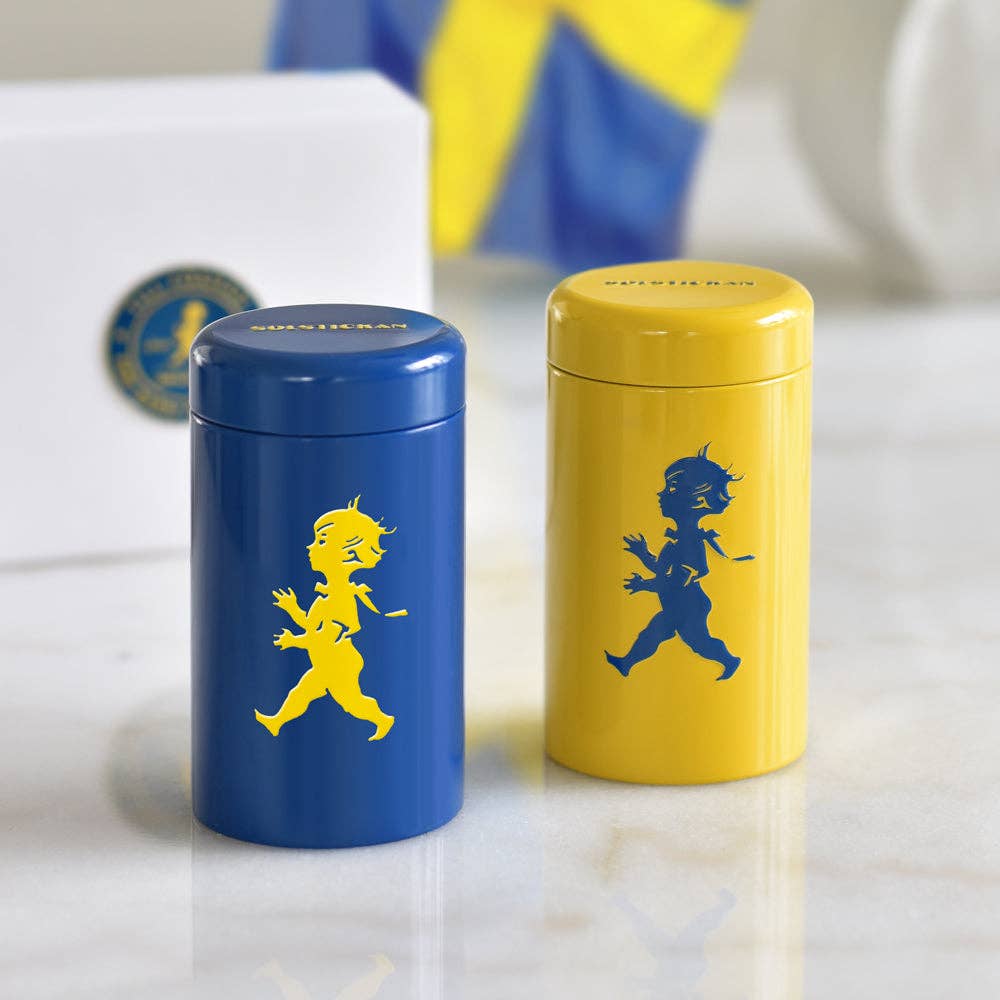 Zwei Solstickan Design Streichholzdosen, eine blaue mit einem gelben Solsticke Boy und eine gelbe (Streichholzdose Gelb), stehen nebeneinander auf Weiß. Eine verschwommene schwedische Flagge unterstreicht ihre schwedische Qualität und extra lange Streichhölzer.