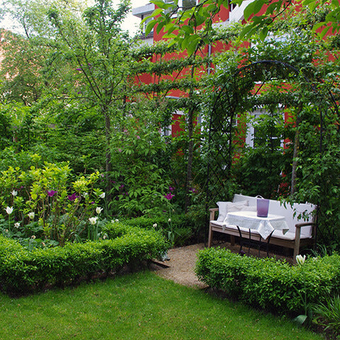 Ein üppiger Garten mit einer Holzbank und einem Tisch unter einem gewölbten Spalier, umgeben von Hecken und Blumen, stand im Mittelpunkt des Kurses am Samstag, 27.2.2027 | 10.00 – 13.00 Uhr: HALBTAGESKURS Grundlagen der Gartengestaltung.