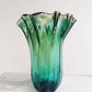 Die große „Fazzoletto“-Vase von Murano Dream aus grünem Murano-Glas (H 32 cm, Ø 24 cm) mit gewelltem Rand steht auf einer weiß-grau gemaserten Fläche vor hellem Hintergrund.