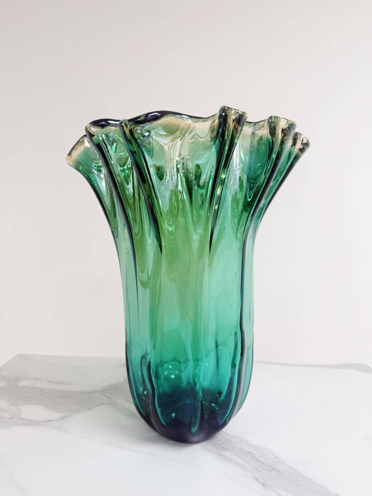Die große „Fazzoletto“-Vase von Murano Dream aus grünem Murano-Glas (H 32 cm, Ø 24 cm) mit gewelltem Rand steht auf einer weiß-grau gemaserten Fläche vor hellem Hintergrund.