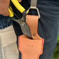 Eine Hand hält eine Gartenschere mit gelbem Griff über einem hellbraunen Niwaki Single Holster mit japanischer Schrift und roten Nähten, das mit dem Niwaki | 'Belt Clip' an einer dunklen Hose befestigt ist. Praktische Halterung für Niwaki Single Holster.