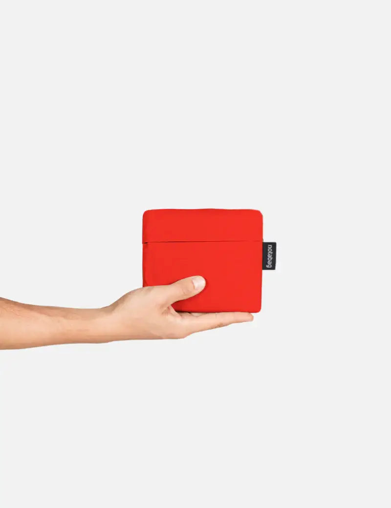 Eine Hand hält eine kompakte rote Notabag | „Original Red“ – Tasche und Rucksack in einem von Notabag mit einem schwarzen Etikett – ideal als wiederverwendbare Einkaufstasche oder praktischer Begleiter für deinen Rucksack und deine Tasche.
