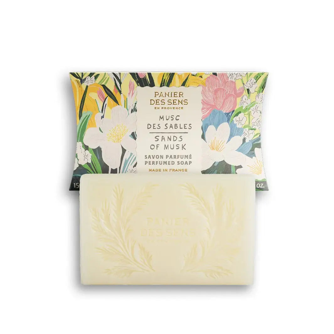 Ein Stück Panier Des Sens | Duftseife Musc des Sables 150 g steht vor seiner blumigen Schachtel, die mit bunten Blumen und grünen Blättern verziert ist. Die Seife ist mit einem eleganten, geprägten botanischen Design versehen.