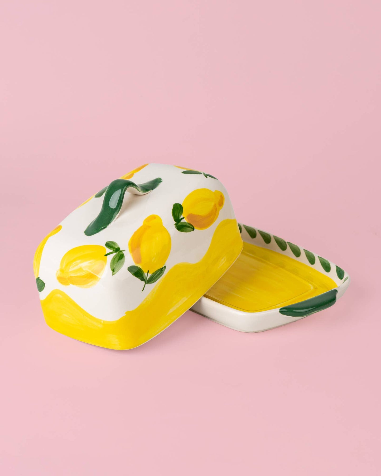 Die handgefertigte Keramik-Butterdose „Limone“ von Alfar Tierra Cocida in Gelb und Weiß präsentiert sich vor einem pinkfarbenen Hintergrund. Der Deckel, kunstvoll mit leuchtenden Zitronen und grünen Blättern verziert, hebt sich leicht von dem gelb-weißen Untergrund ab und unterstreicht stilvoll den mediterranen Charme dieses einzigartigen Tischaccessoires.