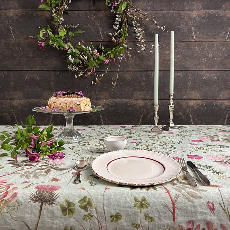 Die Tischdecke „Flora Danica Atlas“ von Koustrupco (350 x 145 cm) schmückt den Tisch, auf dem ein Teller, eine Tasse, Besteck, Kerzen, ein Blätterkranz und eine Torte auf einer Glasplatte stilvoll arrangiert sind – alles vor dem Hintergrund von dunklem Holz.