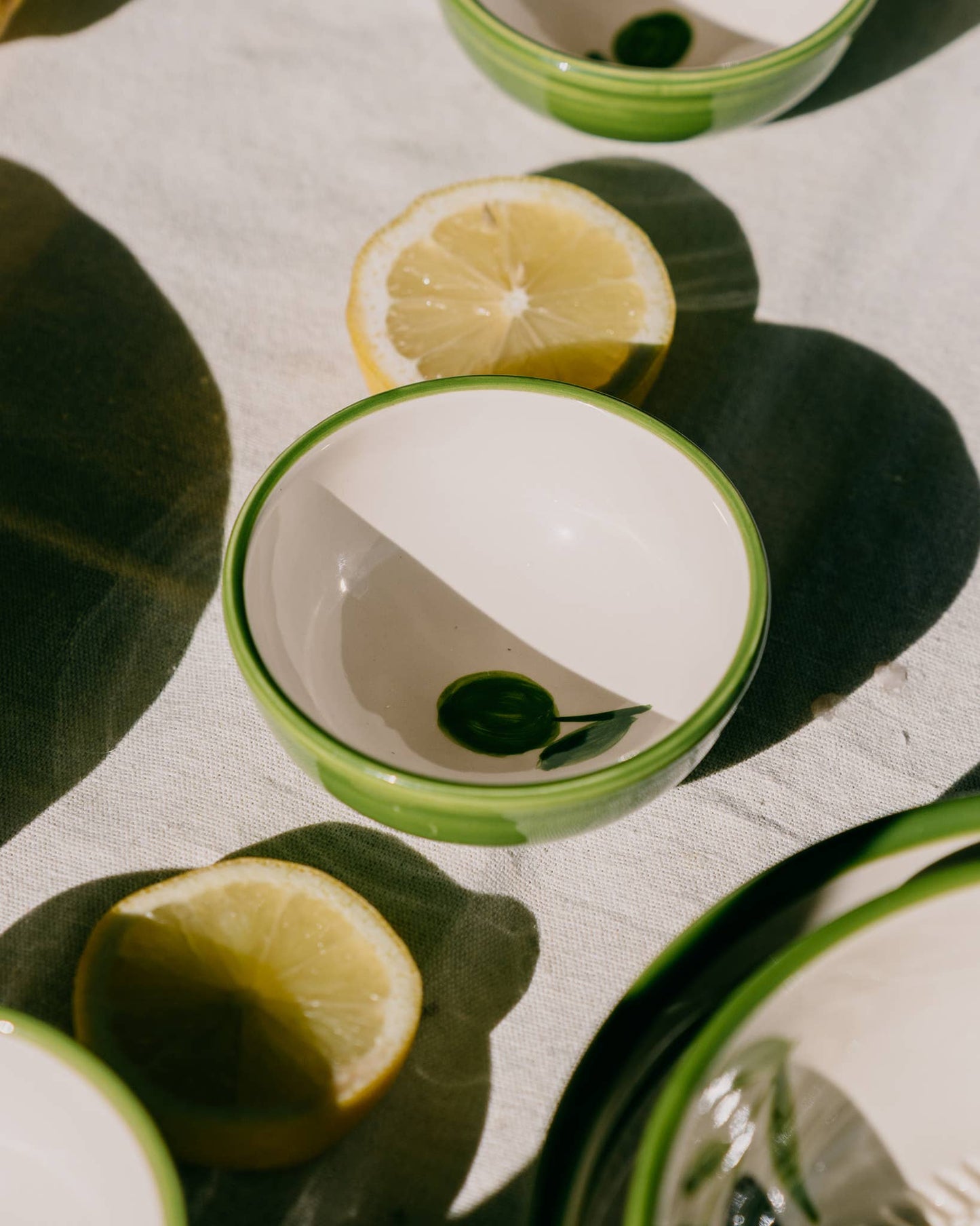 Eine handgefertigte kleine Keramikschale „Olive“ von Alfar Tierra Cocida in Weiß mit grünem Rand steht auf einem Tuch neben Zitronenscheiben und wirft im hellen Sonnenlicht markante Schatten – ein mediterranes Schmuckstück spanischer Handwerkskunst.