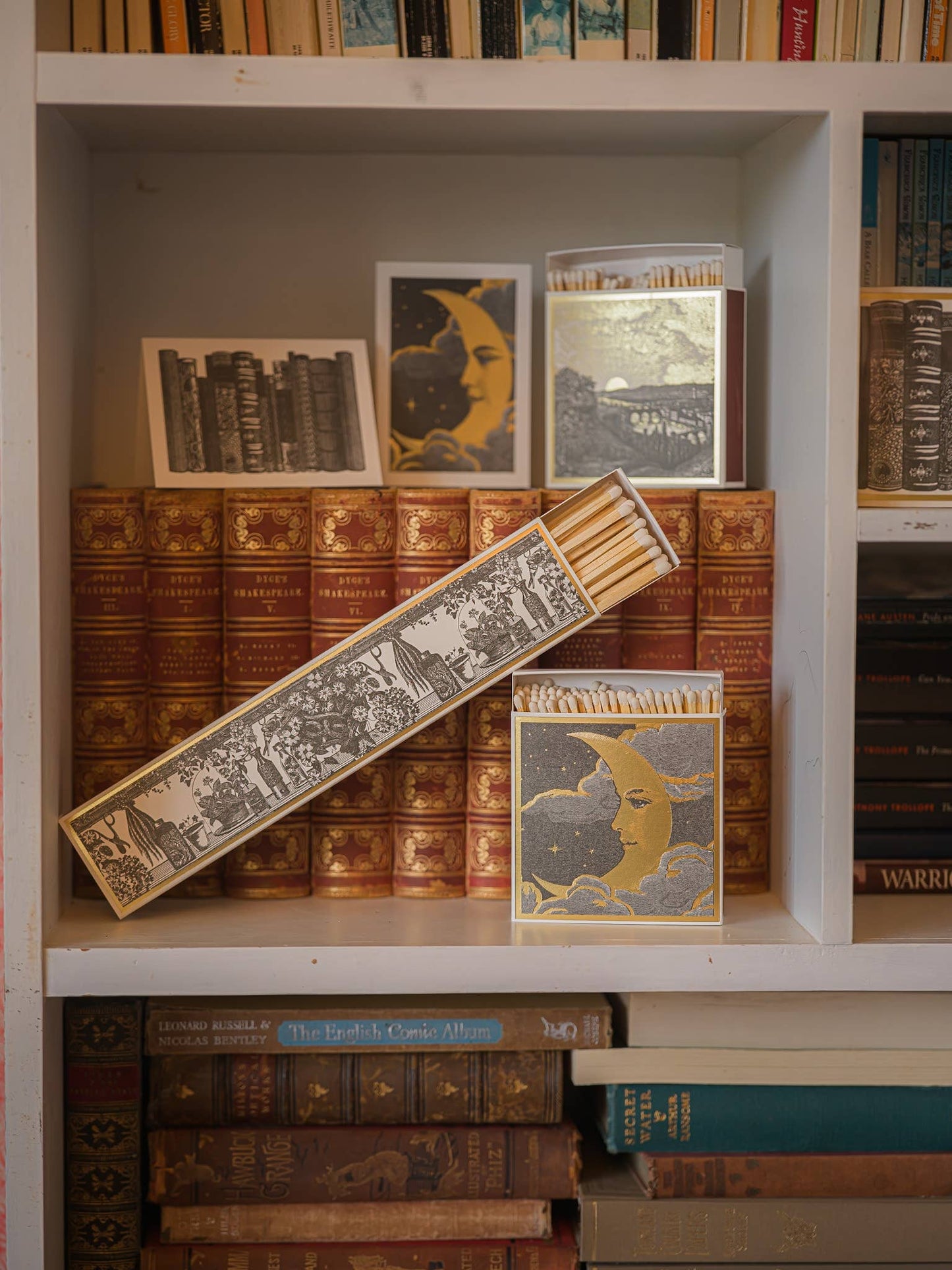 Ein Bücherregal mit alten Büchern und eine geöffnete Schachtel Archivist | 'Land der Träume' Streichhölzer von Archivist Gallery - lange Streichhölzer mit Goldfolienmond und Buchmotiven - sind ein einzigartiges Geschenk für jeden Bücherliebhaber.