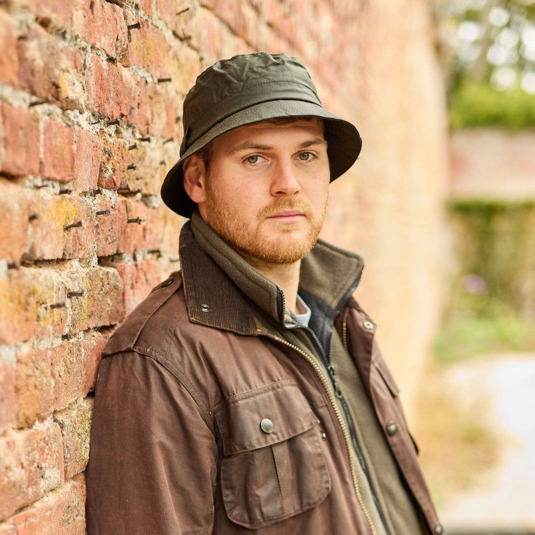 Ein bärtiger Mann, der einen dunklen „Wachstropfen“-Krempenhut von Tweedmill Textiles und eine braune Jacke trägt, lehnt draußen an einer Backsteinmauer und blickt mit ausdruckslosem Gesicht in die Kamera.