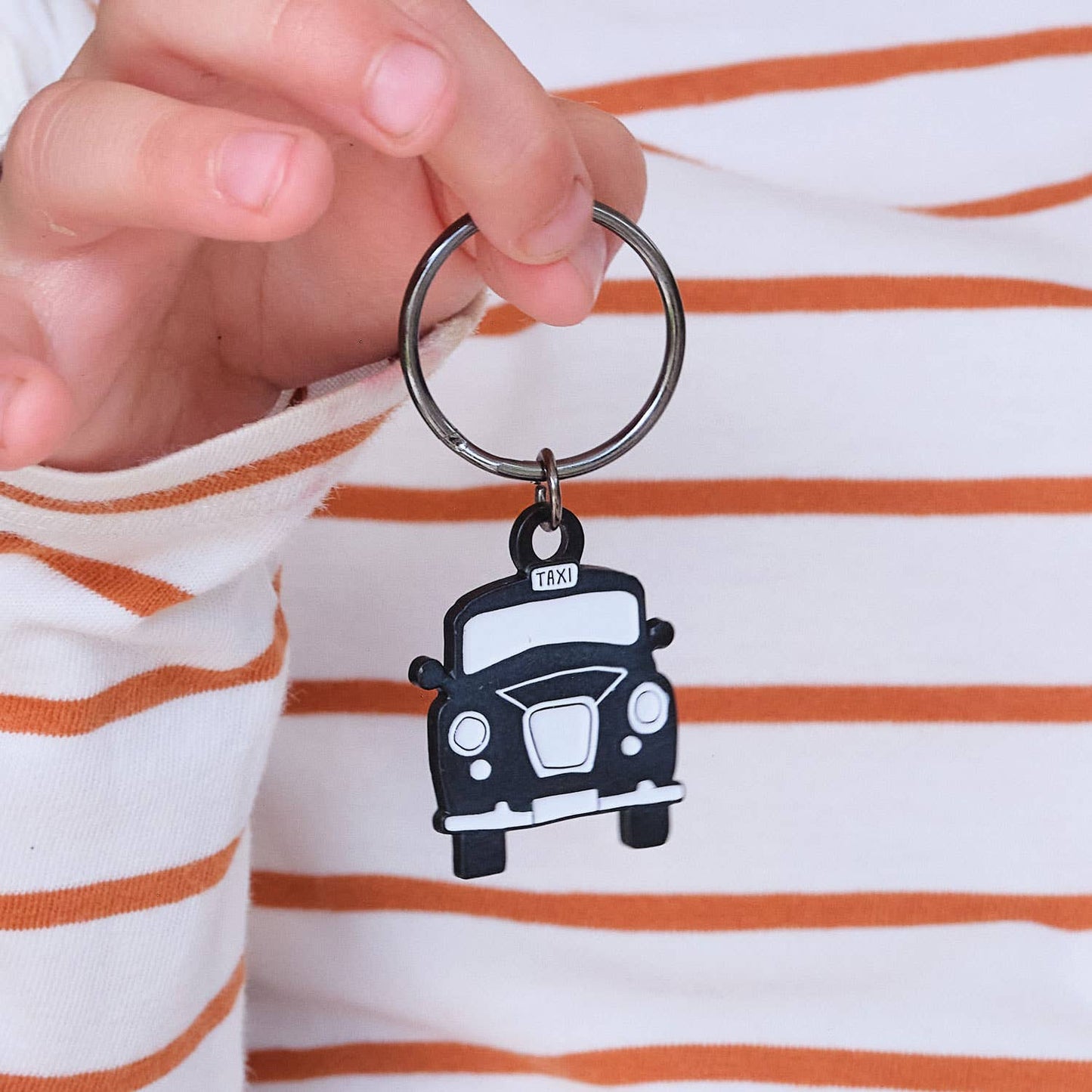 Eine Hand hält den Victoria Eggs - Schwarzen Taxi-Schlüsselanhänger. Die Person trägt ein weißes Shirt mit orangefarbenen Streifen - das ideale Geschenk für London-Fans von Victoria Eggs.