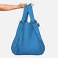 Eine Hand hält die Notabag „Original Blue“ – eine Kombination aus Tasche, Rucksack und Handtasche von Notabag – vor einem schlichten weißen Hintergrund. Die minimalistische blaue Tasche hat ein schwarzes Seitenetikett und eignet sich perfekt für den Alltag.
