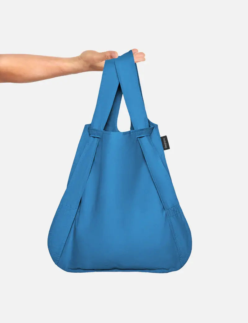 Eine Hand hält die Notabag „Original Blue“ – eine Kombination aus Tasche, Rucksack und Handtasche von Notabag – vor einem schlichten weißen Hintergrund. Die minimalistische blaue Tasche hat ein schwarzes Seitenetikett und eignet sich perfekt für den Alltag.
