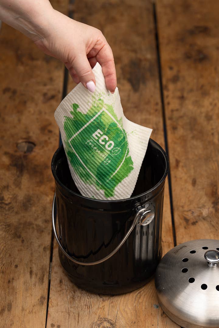 Eine Hand legt das More Joy Home 'Kranz aus Stiefmütterchen' biologisch abbaubares Schwammtuch mit ECO-Label in eine schwarze Komposttonne auf Holz - ein einfacher Schritt zu einem umweltfreundlichen Zero-Waste-Produkt Lifestyle.