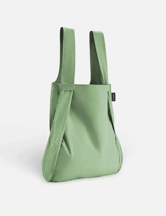 Eine Notabag | Rucksack und Tasche in einem „Olive“ von Notabag mit breiten Tragegriffen steht aufrecht vor einem schlichten weißen Hintergrund. Der weiche und dennoch robuste Stoff sowie ein kleines schwarzes Etikett an einer Seite unterstreichen das stilvolle, wiederverwendbare Design.