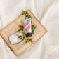 Eine Tube Castelbel | Handcreme Jasmin 60 ml und eine Seifenschale liegen auf einem aufgeschlagenen Buch mit verstreuten grünen Blättern; der blumige Duft schwebt über einem weichen weißen Stoff im Hintergrund.