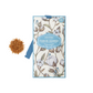 Ein Castelbel | Sachet Cotton Flower 10 g mit floralem Baumwollmuster und blauer Umrandung steht neben braunem Granulat auf weißem Hintergrund.