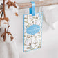 Das Castelbel | Sachet Cotton Flower 10 g hängt mit einem blauen Band an einem Holzstab und verbreitet neben Baumwollzweigen und einem weißen Handtuch lang anhaltenden Duft - ideal als Schrankduft in jedem hellen, luftigen Raum.