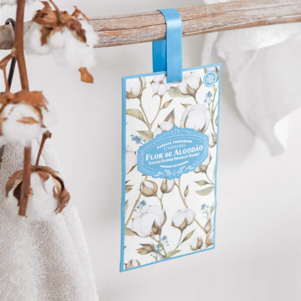Das Castelbel | Sachet Cotton Flower 10 g hängt mit einem blauen Band an einem Holzstab und verbreitet neben Baumwollzweigen und einem weißen Handtuch lang anhaltenden Duft - ideal als Schrankduft in jedem hellen, luftigen Raum.