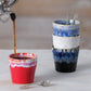 Der Kaffee wird in einen roten Keramikbecher mit weißem und violettem Rand gegossen. Daneben stehen drei gestapelte Costa Nova 'Grespresso' Espressotassen in Denim Blau (9 cl, Steingut und Keramik) mit silbernen Löffeln auf einer hellen Fläche.
