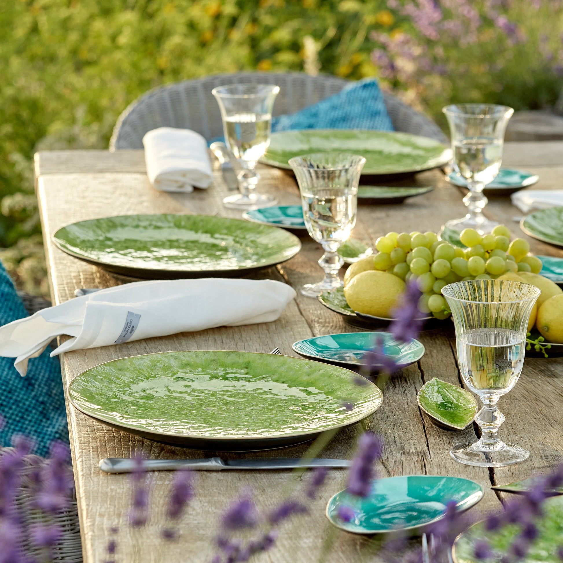 Ein rustikales Tischset für den Außenbereich mit den 11-cm-Tellern "Riviera" von Costa Nova, klaren Gläsern, weißen Servietten, einem Tafelaufsatz aus grünen Trauben und Zitronen und blühenden lila Blumen sorgt für ein mediterranes Ambiente.