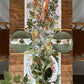 Ein rustikaler Holztisch, gedeckt für zwei Personen, mit grünen Tellern, Servietten mit Blumenmuster, goldenem Besteck, Weingläsern, Kerzen und dem Koustrupco | Tischläufer aus Leinen (145 x 45 cm) im herbstlichen Design als Blickfang sorgt für eine einladende herbstliche Tischdekoration.