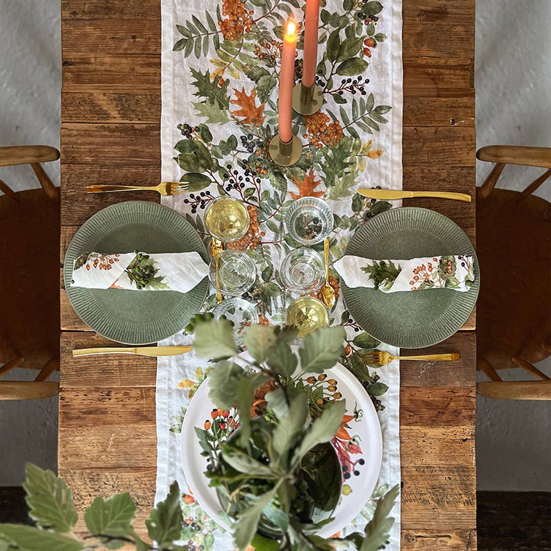 Ein rustikaler Holztisch, gedeckt für zwei Personen, mit grünen Tellern, Servietten mit Blumenmuster, goldenem Besteck, Weingläsern, Kerzen und dem Koustrupco | Tischläufer aus Leinen (145 x 45 cm) im herbstlichen Design als Blickfang sorgt für eine einladende herbstliche Tischdekoration.