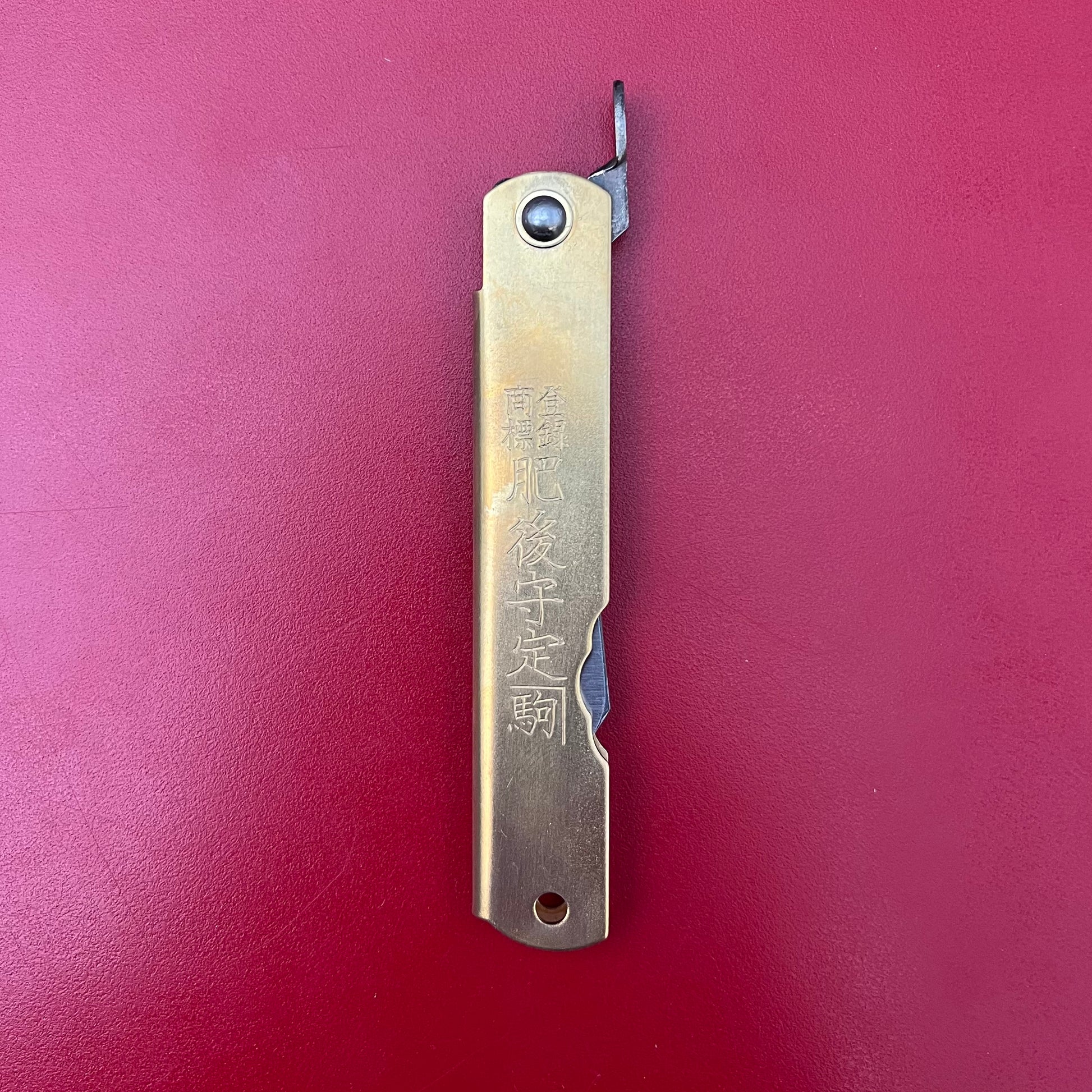Ein Niwaki | 'Blue Steel Higonokami' Klappmesser von Niwaki, mit eingravierten Kanji und einem rechteckigen Messinggriff mit sichtbarer Klingenspitze, ruht geschlossen auf einer flachen roten Oberfläche.