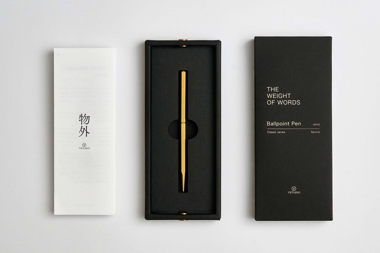 Der ENTREPÔT YSTUDIO Klassischer Revolve Kugelschreiber Schlank, ein eleganter Kugelschreiber aus Goldmessing, wird in einer schwarzen Schachtel zwischen einem weißen japanischen Pamphlet und einer schwarzen Verpackung mit der Aufschrift "THE WEIGHT OF WORDS Ballpoint Pen" präsentiert.