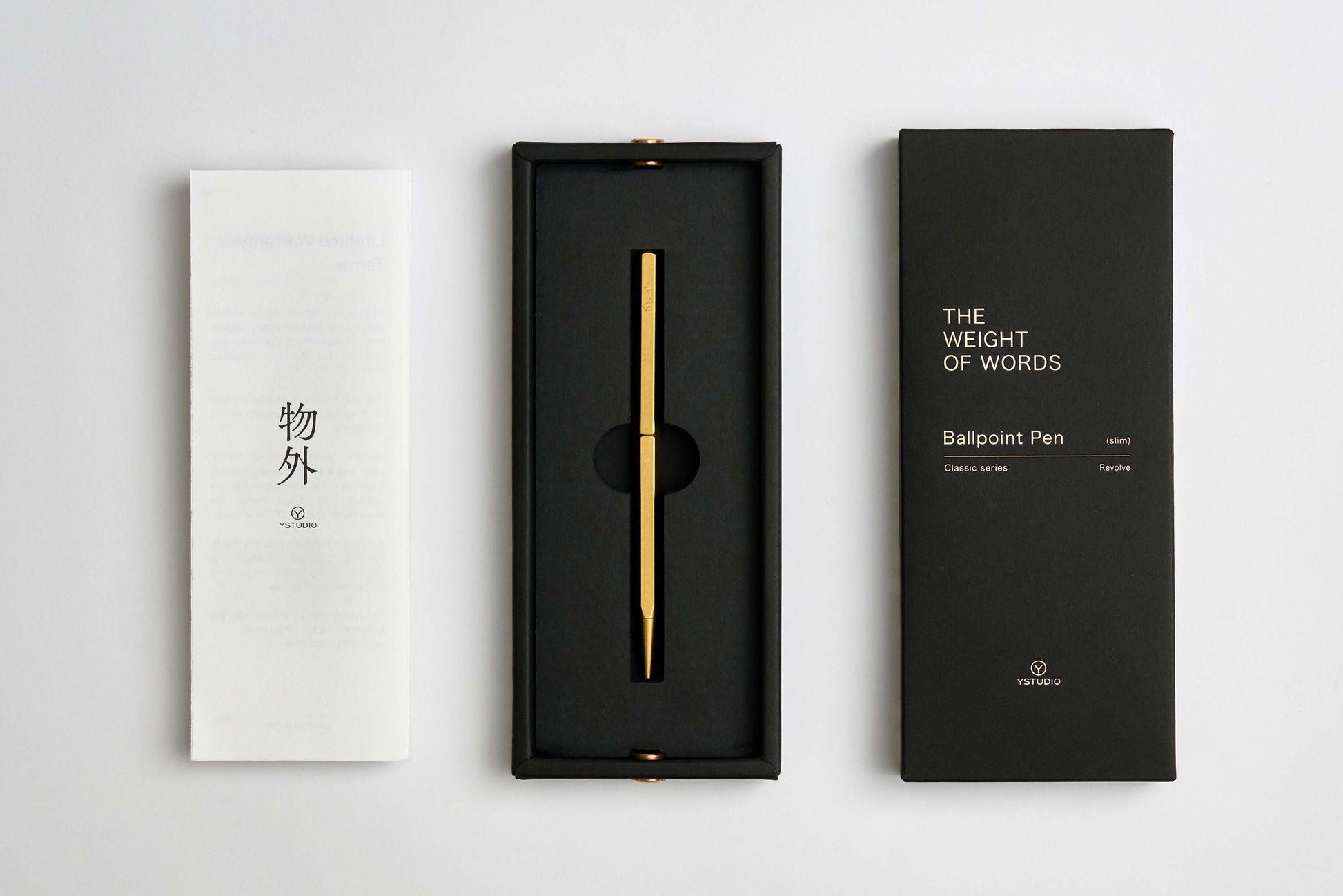 Der ENTREPÔT YSTUDIO Klassischer Revolve Kugelschreiber Schlank, ein eleganter Kugelschreiber aus Goldmessing, wird in einer schwarzen Schachtel zwischen einem weißen japanischen Pamphlet und einer schwarzen Verpackung mit der Aufschrift "THE WEIGHT OF WORDS Ballpoint Pen" präsentiert.