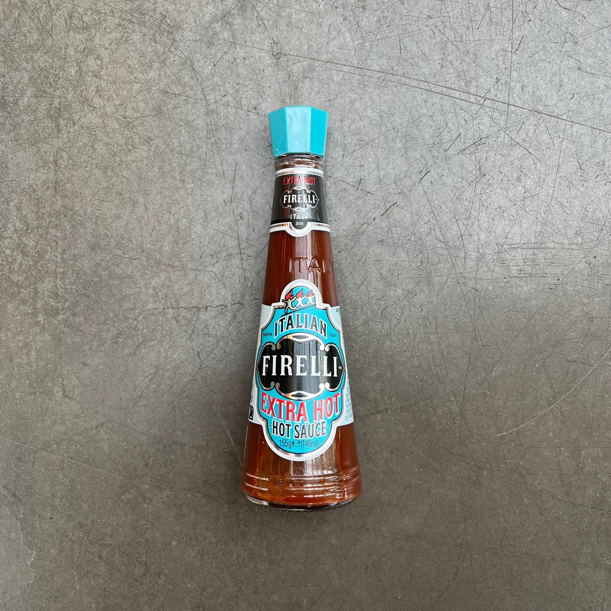Eine Flasche Firelli | Extra scharfe italienische Sauce 148 ml mit blauem Deckel steht auf einer grauen Fläche. Die italienische Sauce von Firelli, mit kalabrischen Chilischoten, passt perfekt zu Grillfleisch. Das Etikett ist blau, schwarz und rot.