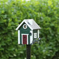Das Green Cottage Nistkasten & Futterhaus von Wildlife Garden steht stilvoll auf einem Pfahl mit weißer Umrandung und roter Tür und ist im skandinavischen Design gehalten. Am Eingang hockt ein Vogel vor einem üppig grünen, blattreichen Hintergrund.