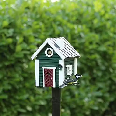 Das Green Cottage Nistkasten & Futterhaus von Wildlife Garden steht stilvoll auf einem Pfahl mit weißer Umrandung und roter Tür und ist im skandinavischen Design gehalten. Am Eingang hockt ein Vogel vor einem üppig grünen, blattreichen Hintergrund.