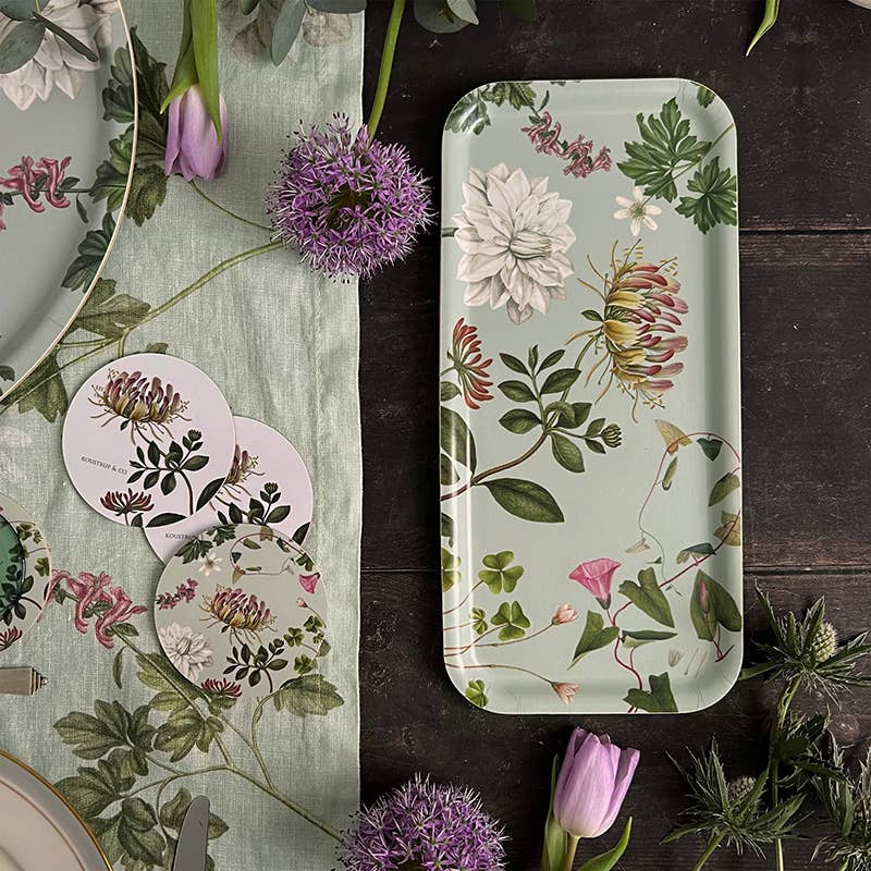 Eine Tischdekoration mit Blumenmotiv, bestehend aus dem Serviertablett „Flora Danica Atlas“ von Koustrupco (32 × 15 cm), passenden Untersetzern, einem Tischläufer mit Blumenmuster, violetten Tulpen und anderen kleinen violetten Blumen, die auf einer dunklen Holzfläche arrangiert sind.
