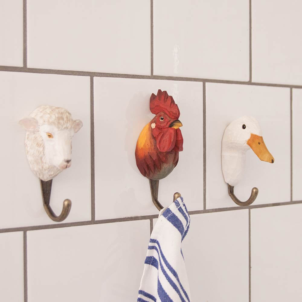 Drei Wandhaken in Form von Tierköpfen - Schaf, Hahn und Ente - sind an einer gefliesten Wand angebracht. Der Wildlife Garden Handgeschnitzter Wandhaken aus Holz 'Hahn' hält ein weißes Handtuch mit blauen Streifen.