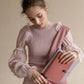 Eine junge Frau mit lockigem Haar und rosa Rüschenbluse packt ihr Tinne Mia Notizbuch mit Knopf 'Leinen Rose Dawn' in eine passende rosa Tragetasche und konzentriert sich auf ihre Aufgabe in einer gedämpften, neutralen Umgebung.