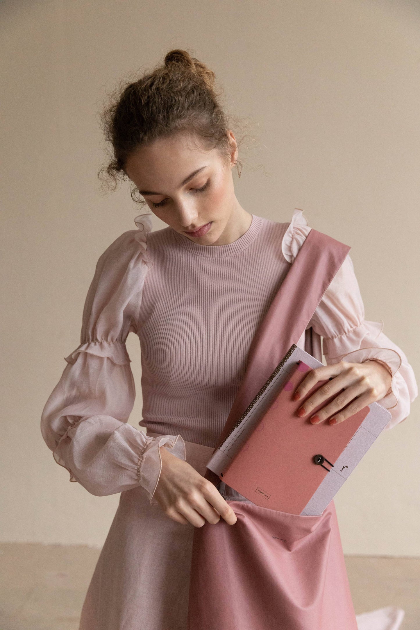 Eine junge Frau mit lockigem Haar und rosa Rüschenbluse packt ihr Tinne Mia Notizbuch mit Knopf 'Leinen Rose Dawn' in eine passende rosa Tragetasche und konzentriert sich auf ihre Aufgabe in einer gedämpften, neutralen Umgebung.