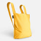 Der Notabag „Original Golden“ von Notabag ist eine leuchtend gelbe, umweltfreundliche Kombination aus Tasche und Rucksack mit zwei Schulterriemen und minimalistischem Design vor hellem Hintergrund.